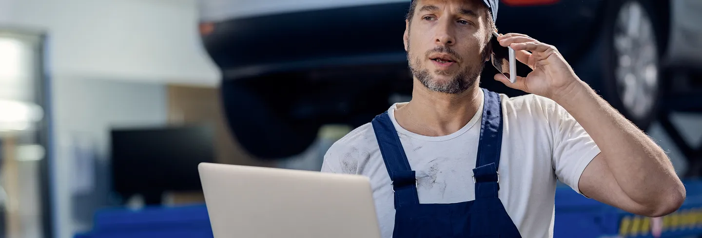 Mechanic wearing a white shirt and blue overalls talking on a smartphone while holding a laptop in a garage.