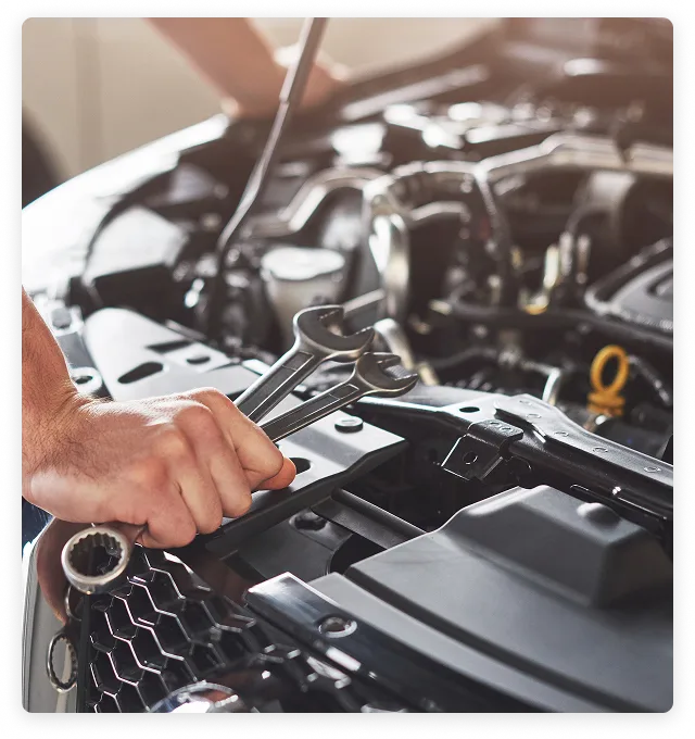 Close-up of a mechanic's hand holding two wrenches while working on a car engine under the open hood.